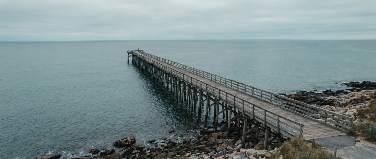 Aerial view of a long wooden pier extending into calm grey-blue Atlantic water, a strong leading line toward the horizon